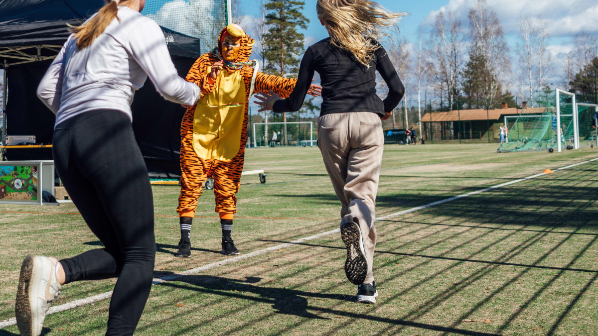 Kaksi nuorta juoksee urheilukentällä ja antaa läpsyn tiikeripukuiselle henkilölle. Kuva: Essi Kultanen.