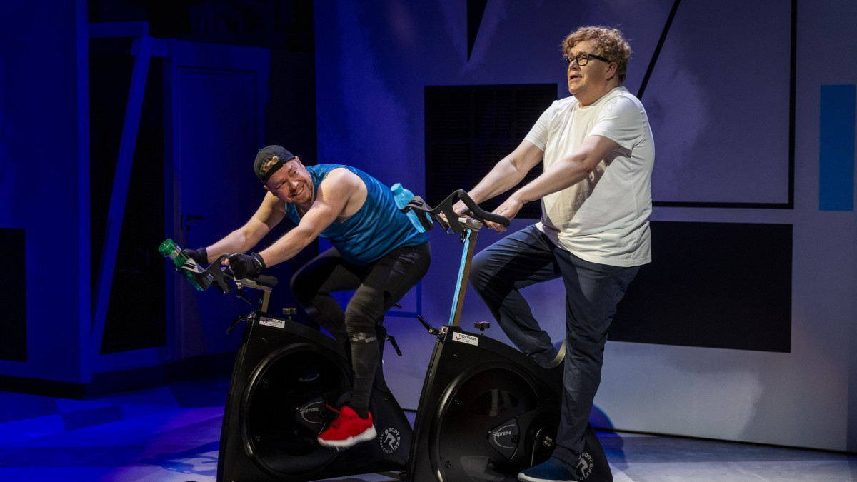 Kaksi miesnäyttelijää polkee lavalla kuntopyöriä.