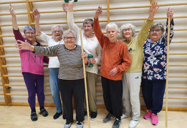 Voimaa vanhuuteen -ryhmän seitsemän naista nostaa käden ilmaan puolapuiden edessä.